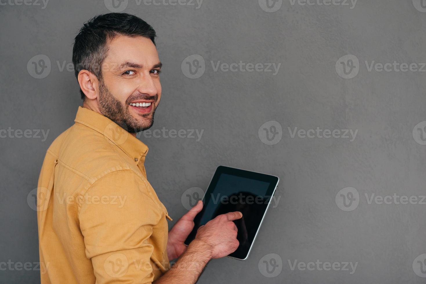 How may I help you  Rear view of handsome mature man holding digital tablet and looking over shoulder with smile while standing against grey background photo