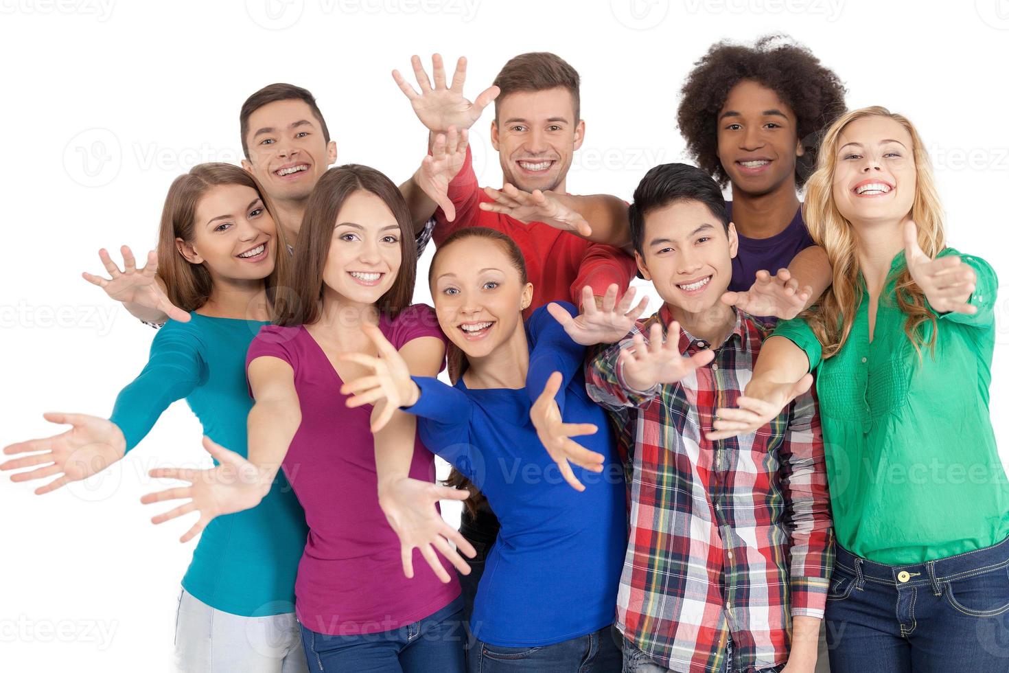 Únase a nosotros, un grupo de alegres jóvenes multiétnicos parados uno cerca del otro y gesticulando mientras están aislados en blanco foto