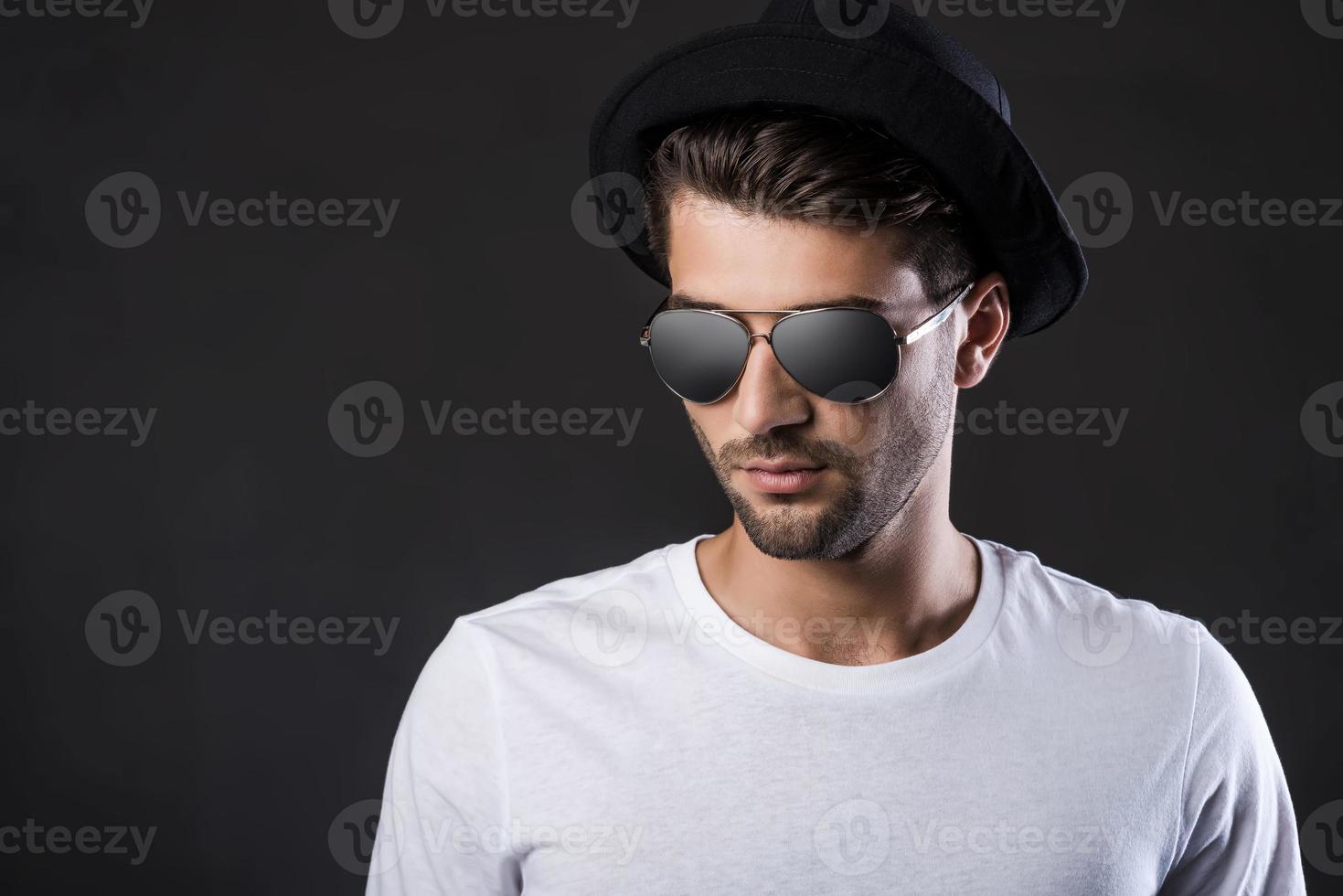 confiado y guapo. apuesto joven con gafas de sol y fedora de pie contra el fondo negro foto