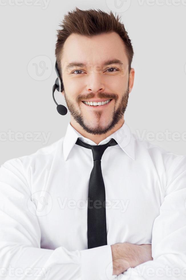 Confident male operator. Handsome young man in formalwear and headset looking at camera and smiling while standing against grey background photo