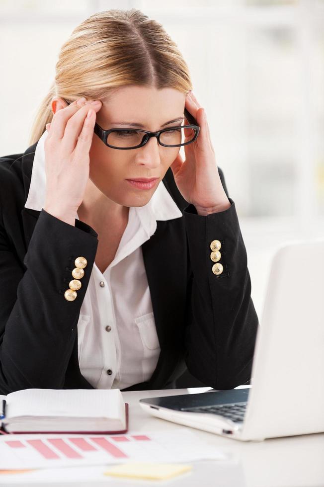 Feeling headache.  Depressed mature woman in formalwear holding head in hands and looking at the laptop while sitting at her working place photo