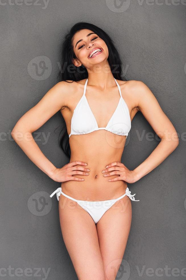 sintiéndose genial y luciendo hermosa. hermosa joven mujer de raza mixta en bikini blanco cogido de la mano en la cadera y sonriendo mientras está de pie contra el fondo gris foto