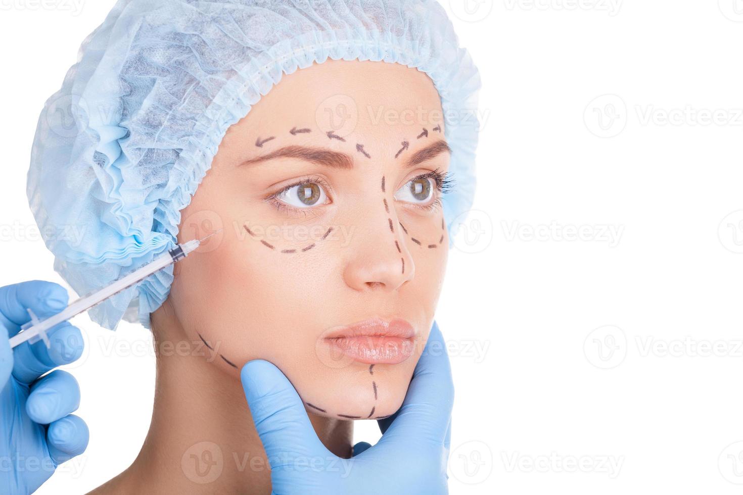 inyección de bótox. hermosa mujer joven con sombreros médicos y bocetos en la cara mirando hacia otro lado mientras los médicos hacen una inyección en la cara foto