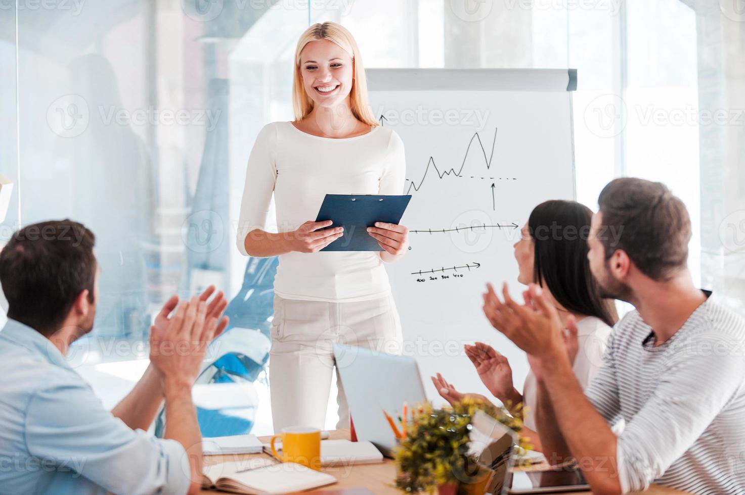 Great presentation Cheerful young woman standing near whiteboard and smiling while her colleagues sitting at the desk and applauding photo