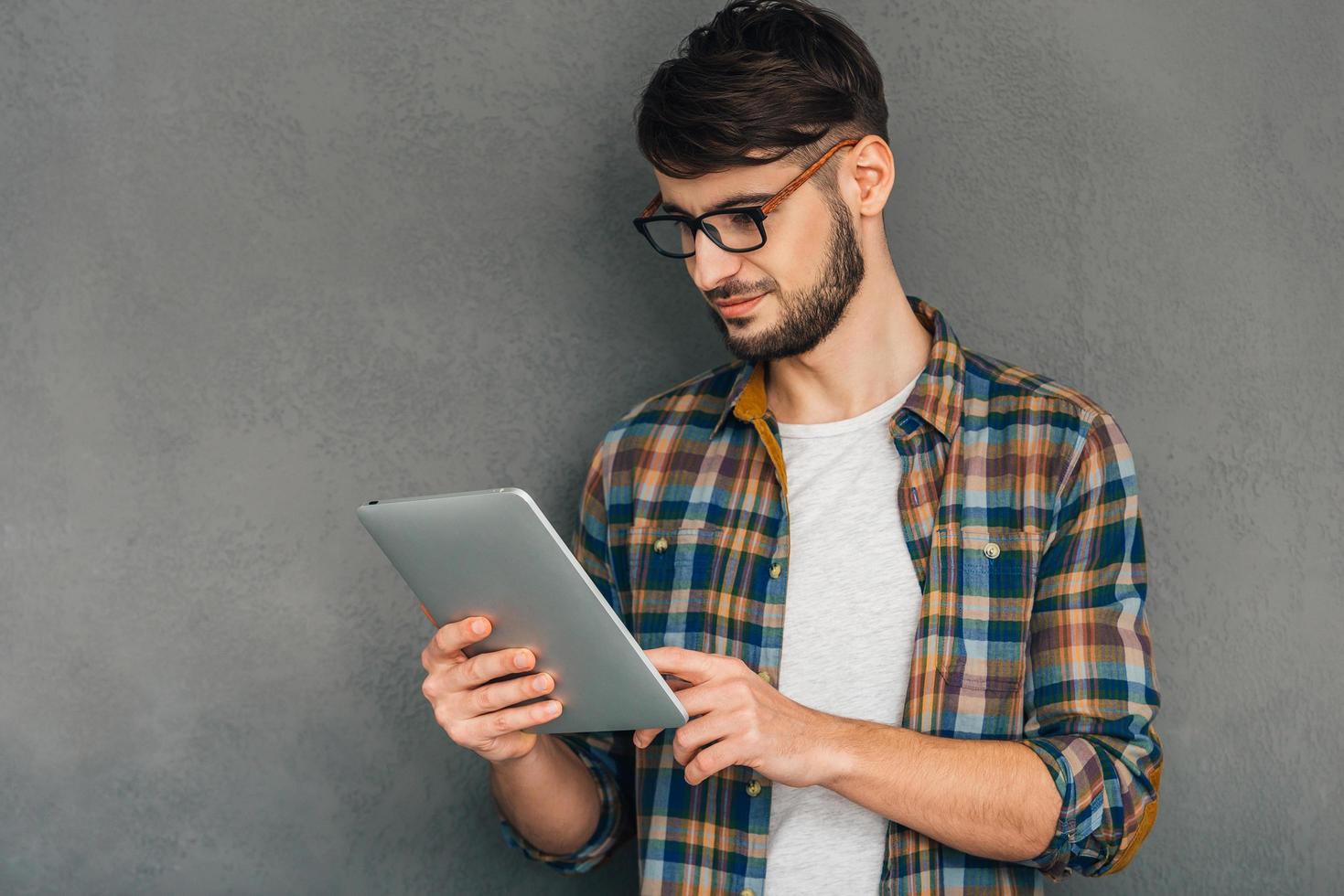 Enjoying his digital tablet. Confident young man holding his digital tablet and looking at it while standing against grey background photo