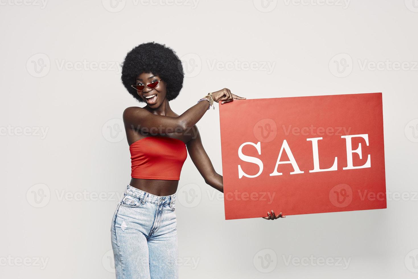 bella joven africana sosteniendo un cartel de venta y sonriendo mientras se enfrenta a un fondo gris foto
