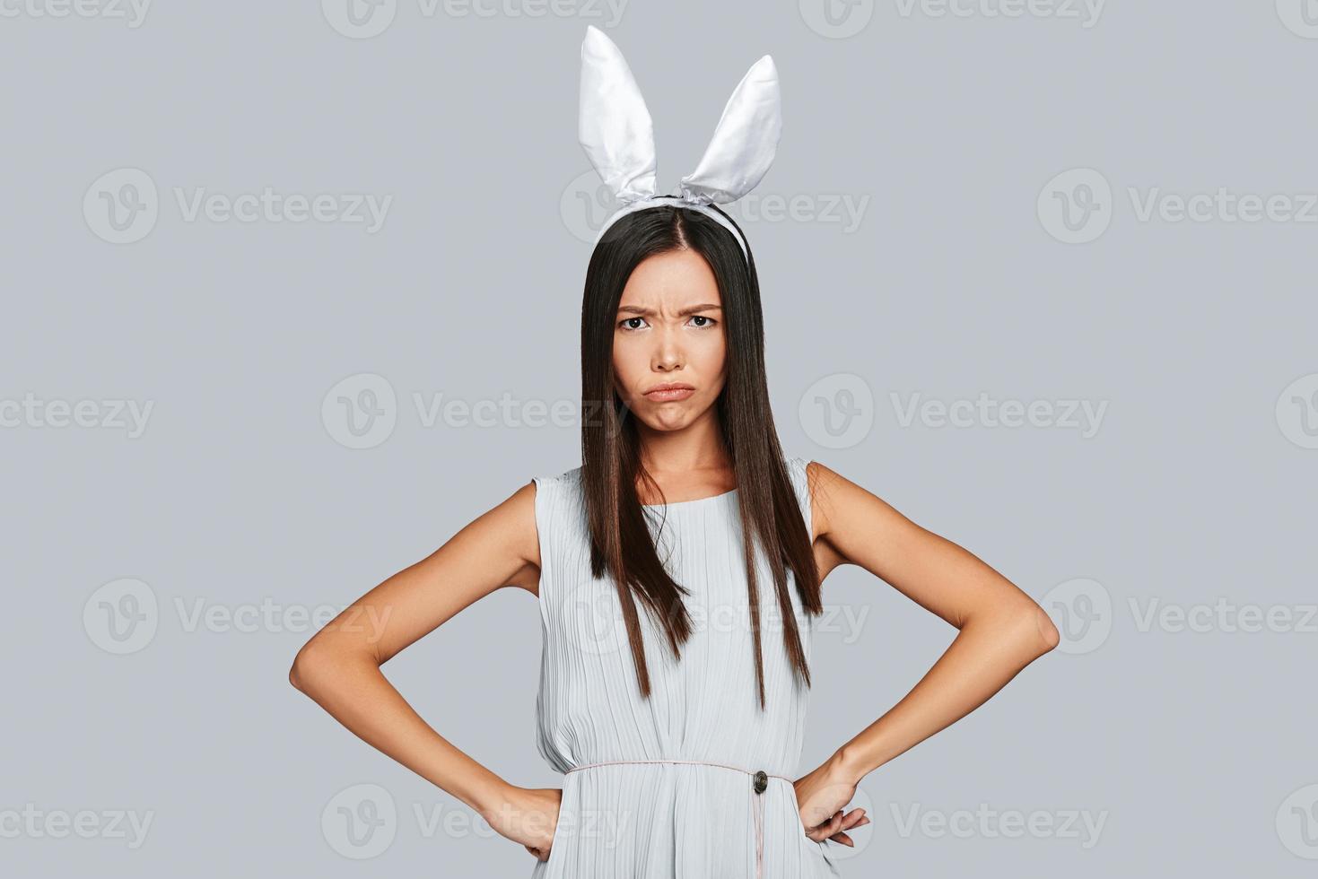 Bored bunny. Angry young Asian woman in bunny ears looking at camera while standing against grey background photo