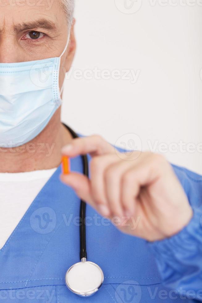 Take this pill Cropped image of doctor in surgical mask showing a pill and looking at camera while standing isolated on white photo