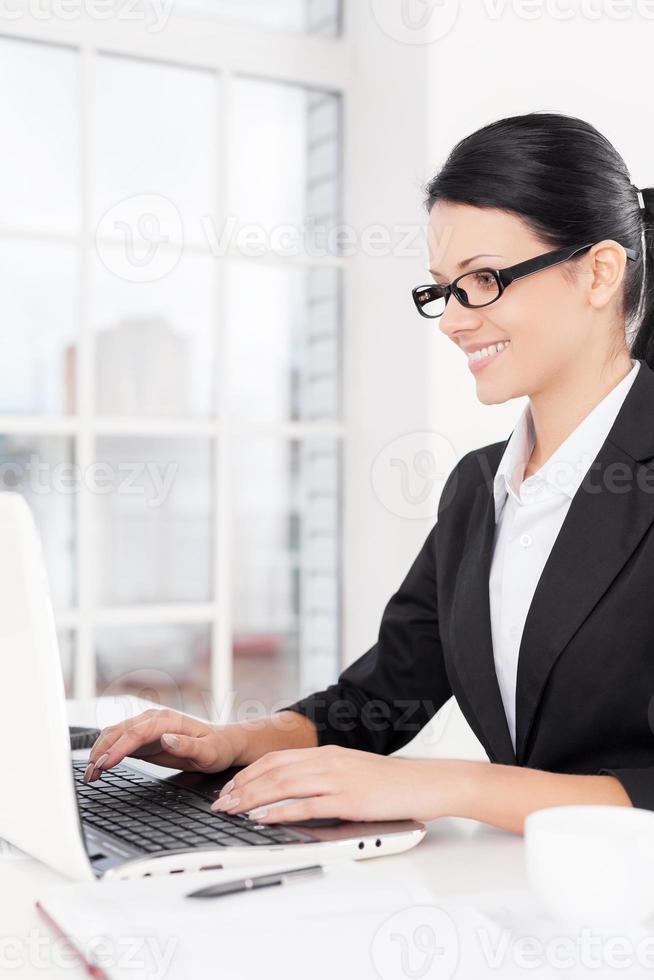 empresaria en el trabajo. vista lateral de una joven mujer de negocios segura de sí misma que usa computadora y sonríe mientras está sentada en su lugar de trabajo foto