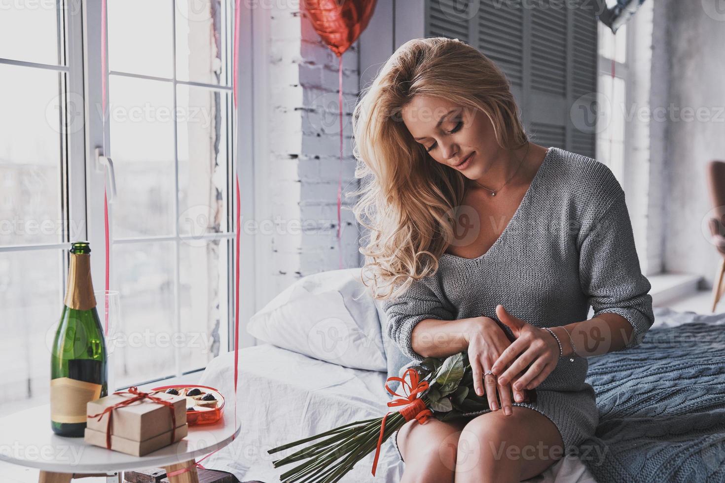 He finally put the question. Attractive young woman touching her engagement ring and holding a bouquet of red roses while sitting on the bed at home photo