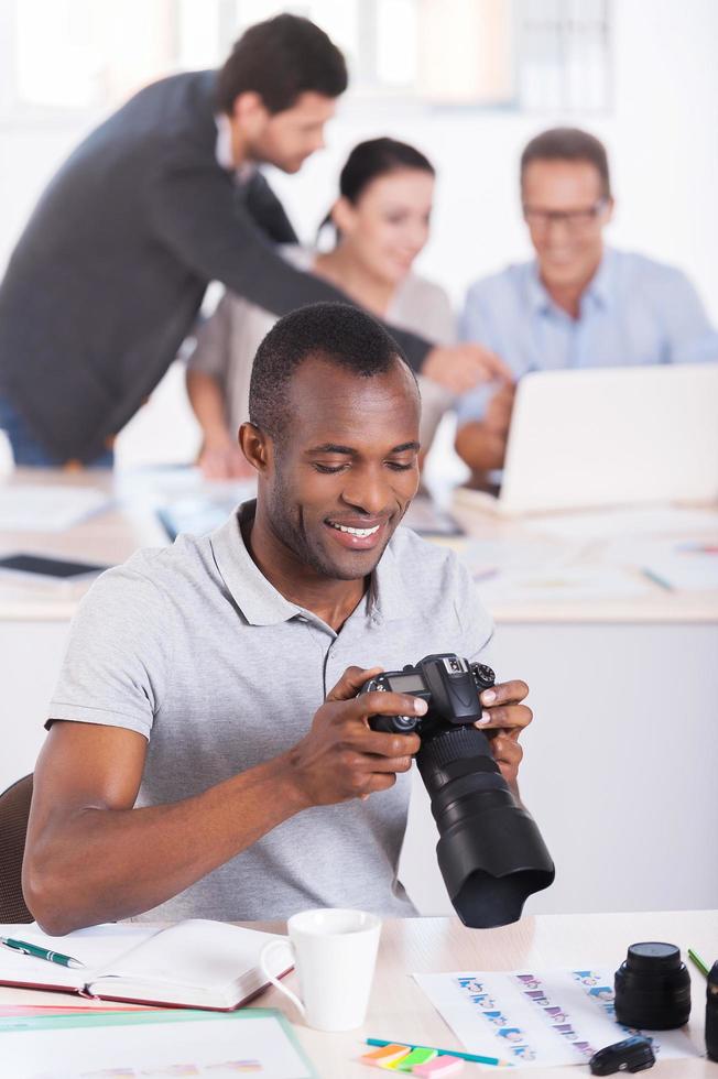 Confident in his work. Confident young African man holding camera and smiling while three people working on background photo