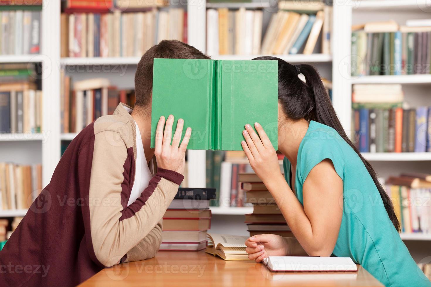 Library romance. Young man and woman sitting close to each other at the library desk and hiding their faces behind a book photo