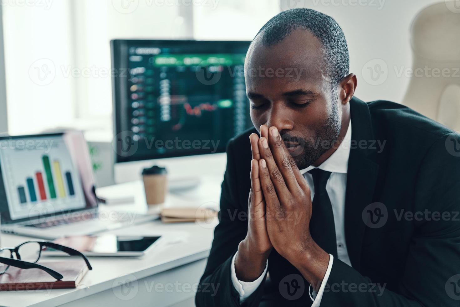 Tired young African man in formalwear keeping hands clasped while working in the office photo
