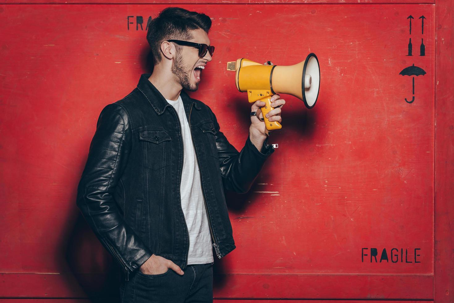 Fashion announcement. Side view of handsome young man in sunglasses holding megaphone and keeping mouth open while standing against red background photo