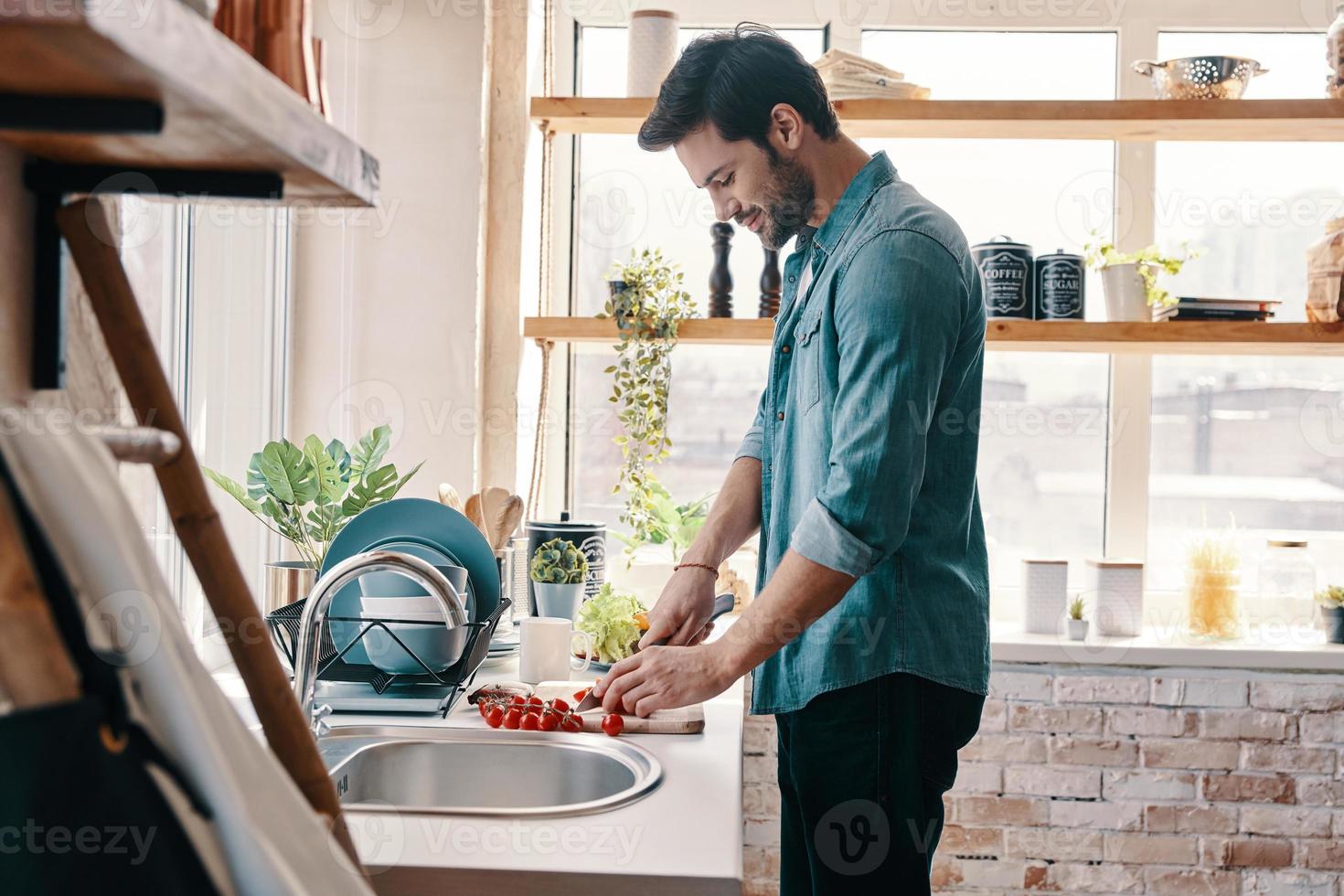 Time to have breakfast. Handsome young man in casual wear chopping food and smiling while standing in the kitchen at home photo