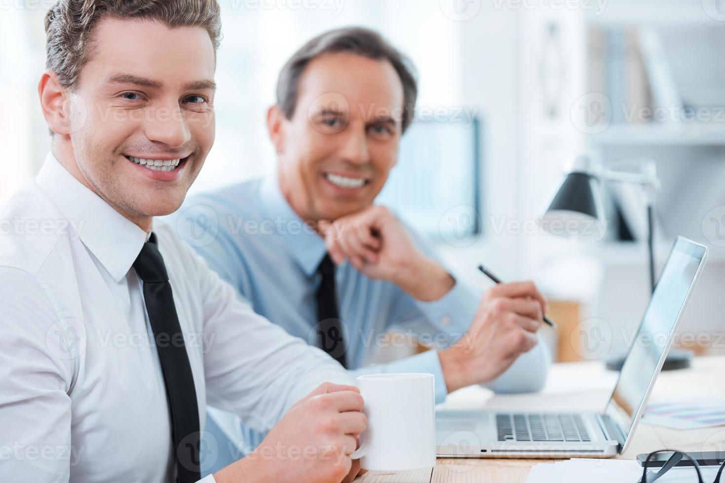 Business partners at work. Side view of two cheerful business people in formalwear smiling at camera while sitting at working place photo