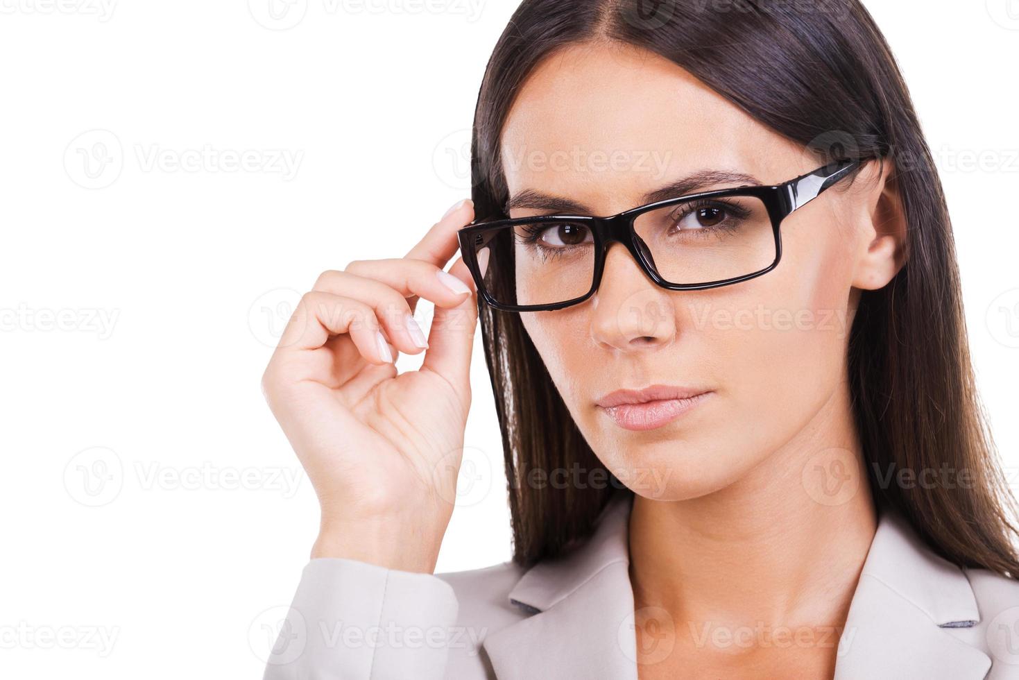 How may I help you Beautiful young businesswoman in suit adjusting her eyeglasses and looking at camera while standing against white background photo