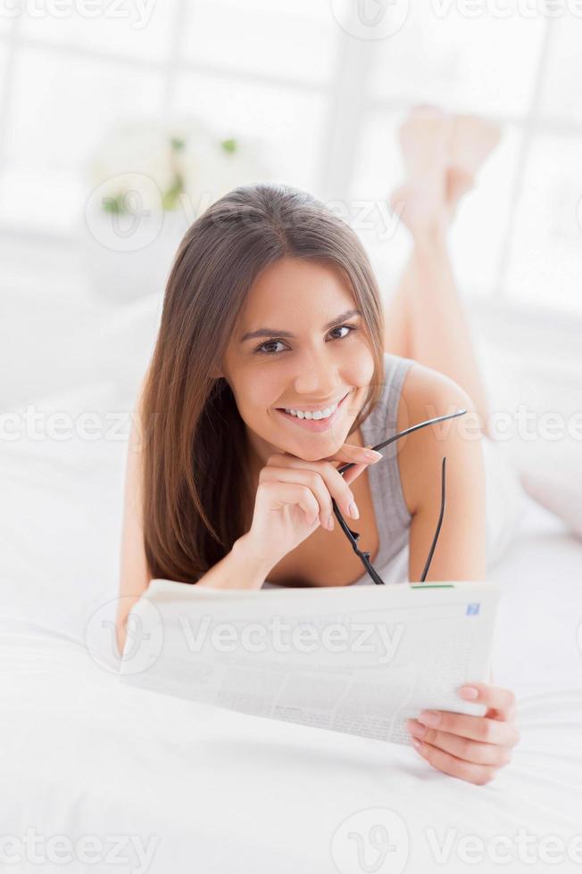 leyendo un periódico de la mañana. bella joven sonriente acostada en la cama y leyendo un periódico foto