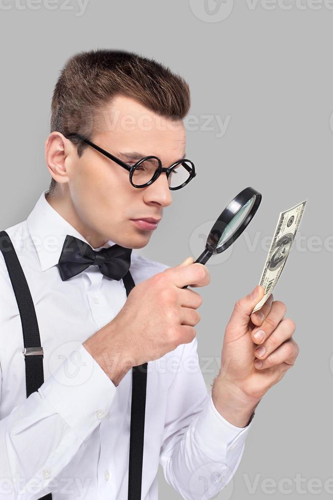 Fake or real Portrait of cheerful young man in bow tie and suspenders looking through magnifying glass at the dollar bill while standing against grey background photo