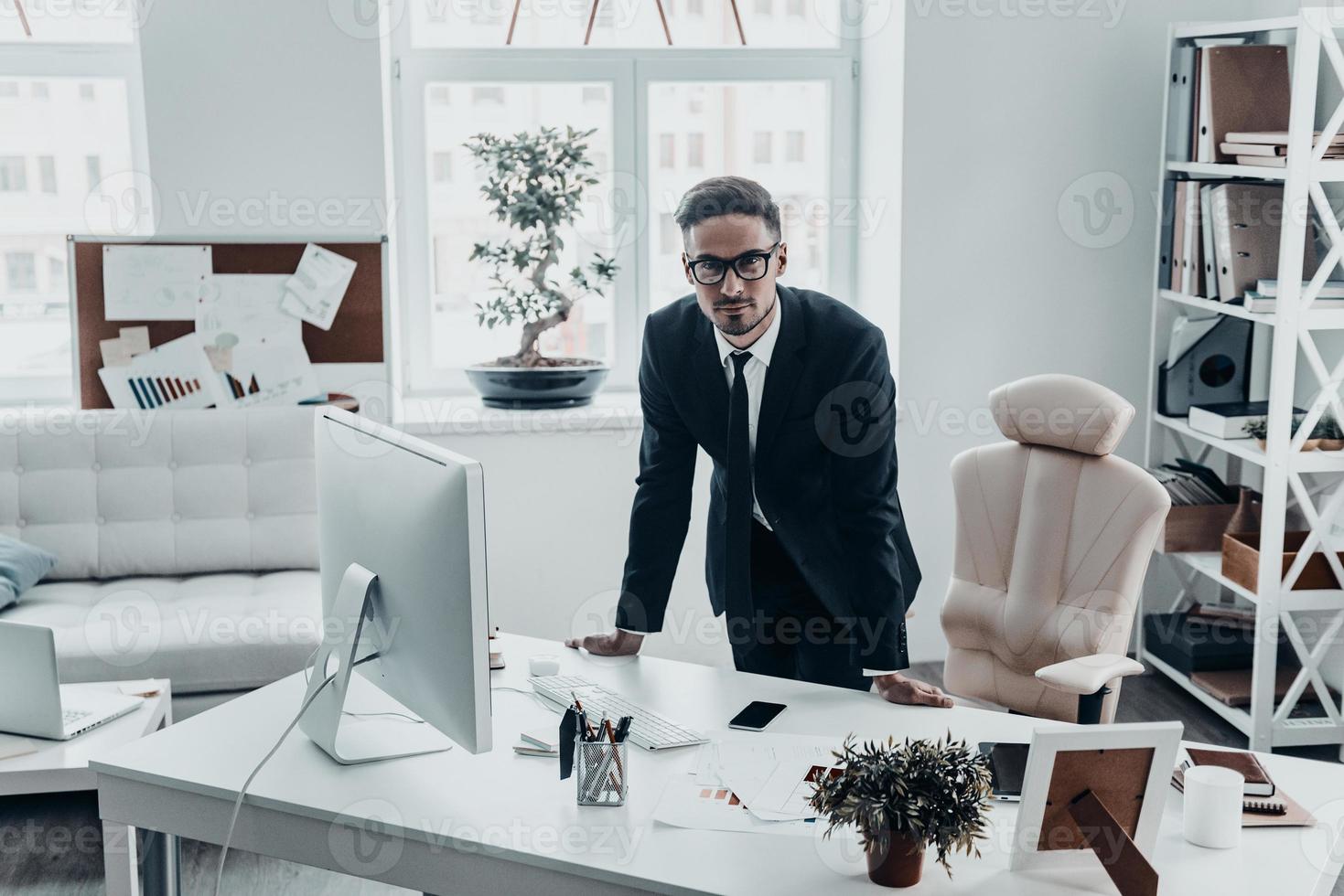 May I help you Handsome young man in full suit looking at camera with serious face while leaning on the office desk photo
