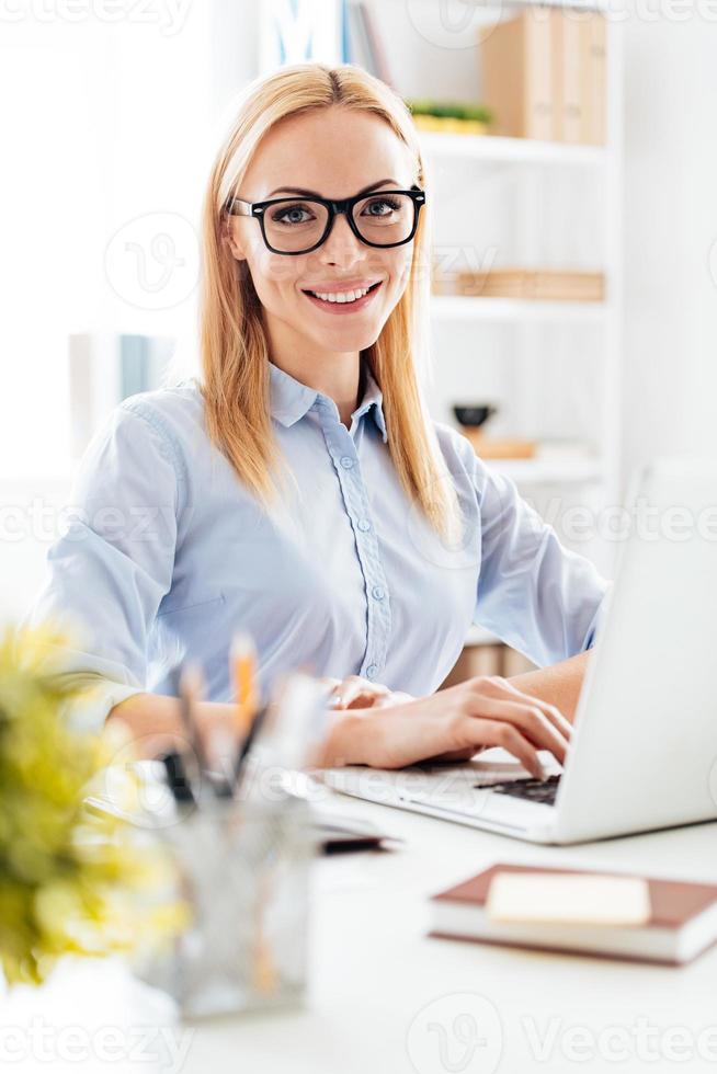 Elegant and cheerful businesswoman. Cheerful young beautiful woman in glasses looking at camera with smile while sitting at her working place photo