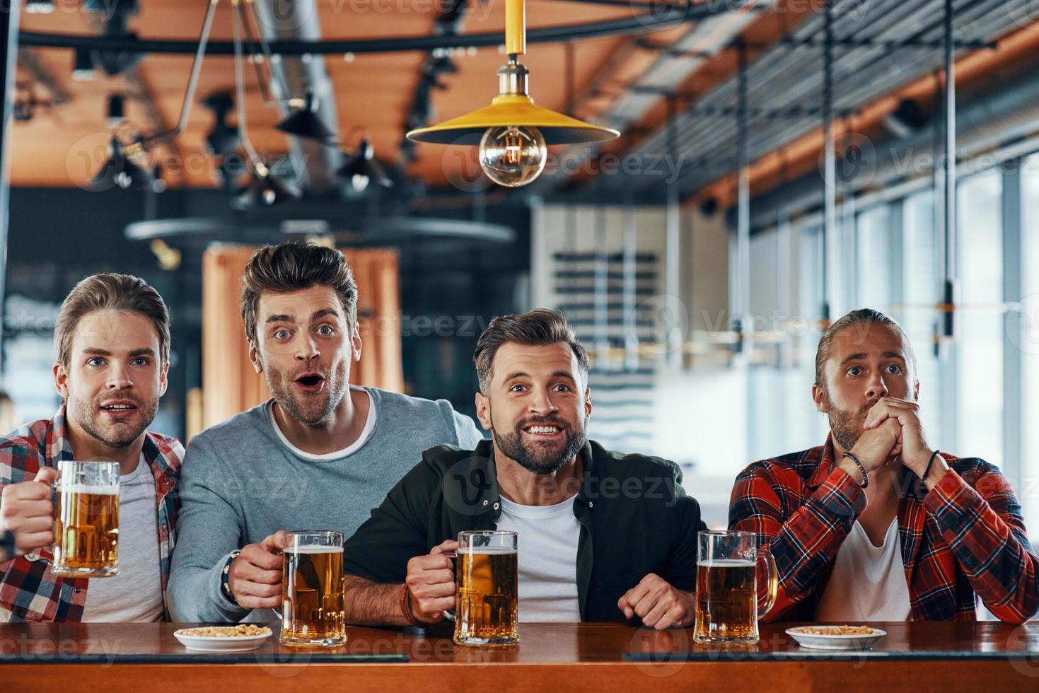 Cheering young men in casual clothing drinking beer and watching sport game while sitting in the pub photo