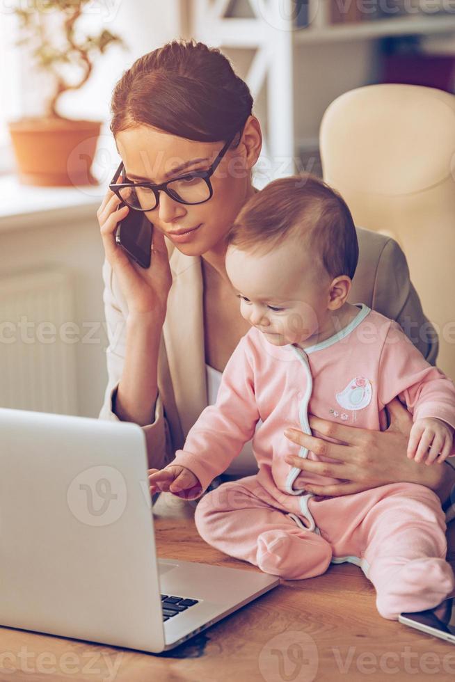 Working with baby. Young beautiful businesswoman talking on mobile phone and looking at laptop while sitting with her baby girl at her working place photo