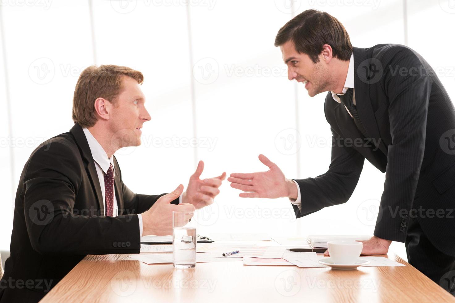 conflicto empresarial. dos jóvenes en ropa formal discutiendo y gesticulando mientras están sentados en la mesa foto