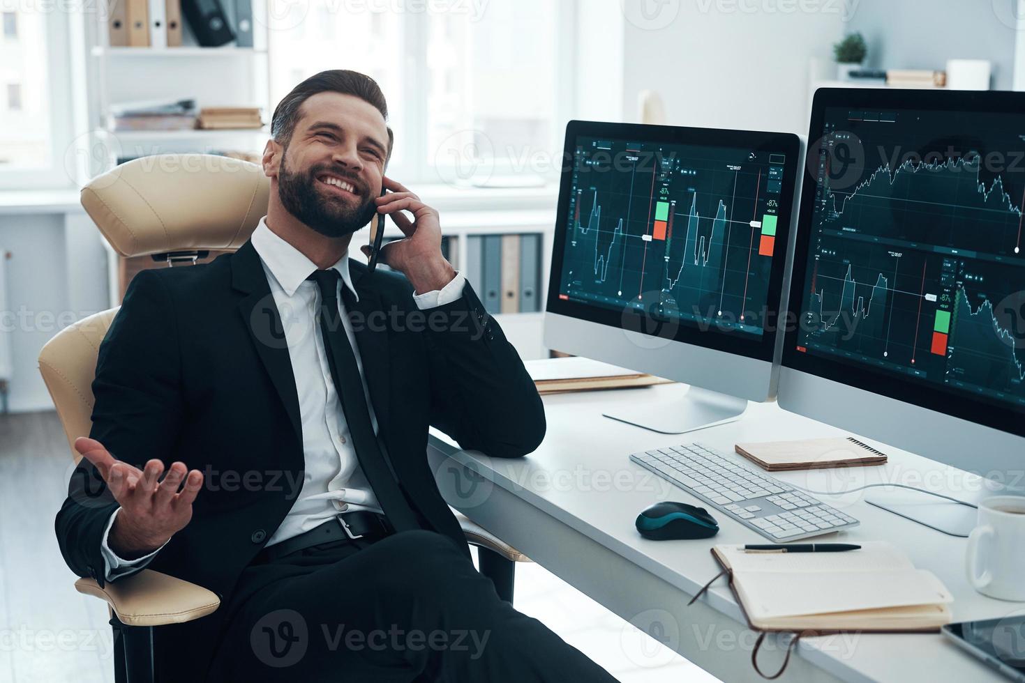 apuesto joven con camisa y corbata hablando por teléfono y sonriendo mientras trabaja en la oficina foto
