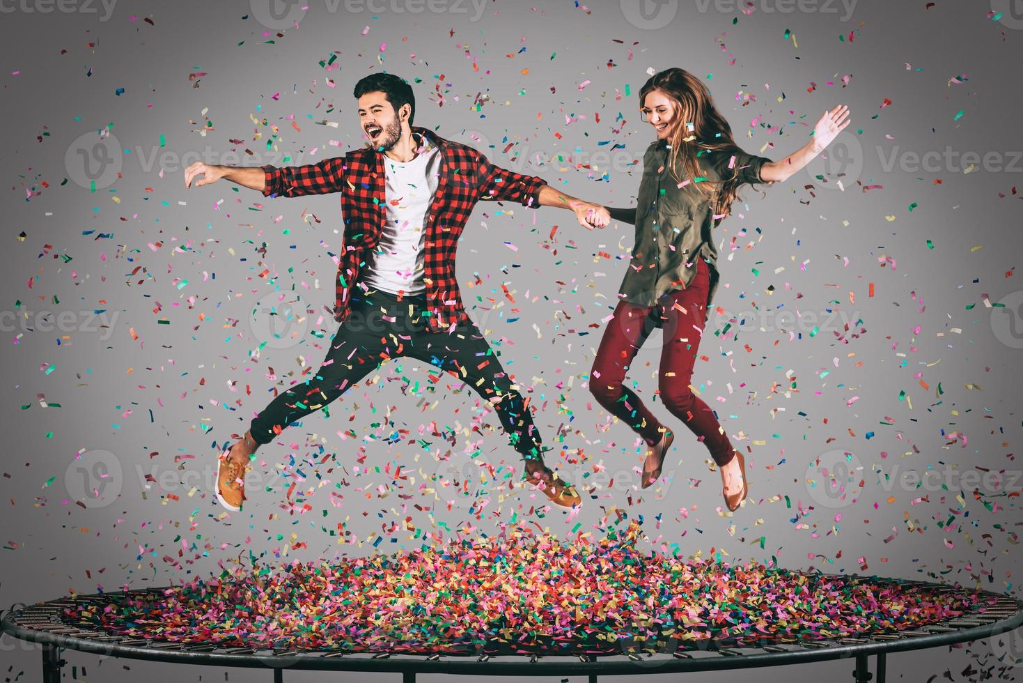 Enjoying fun time together. Mid-air shot of beautiful young cheerful couple holding hands while jumping on trampoline together with confetti all around them photo