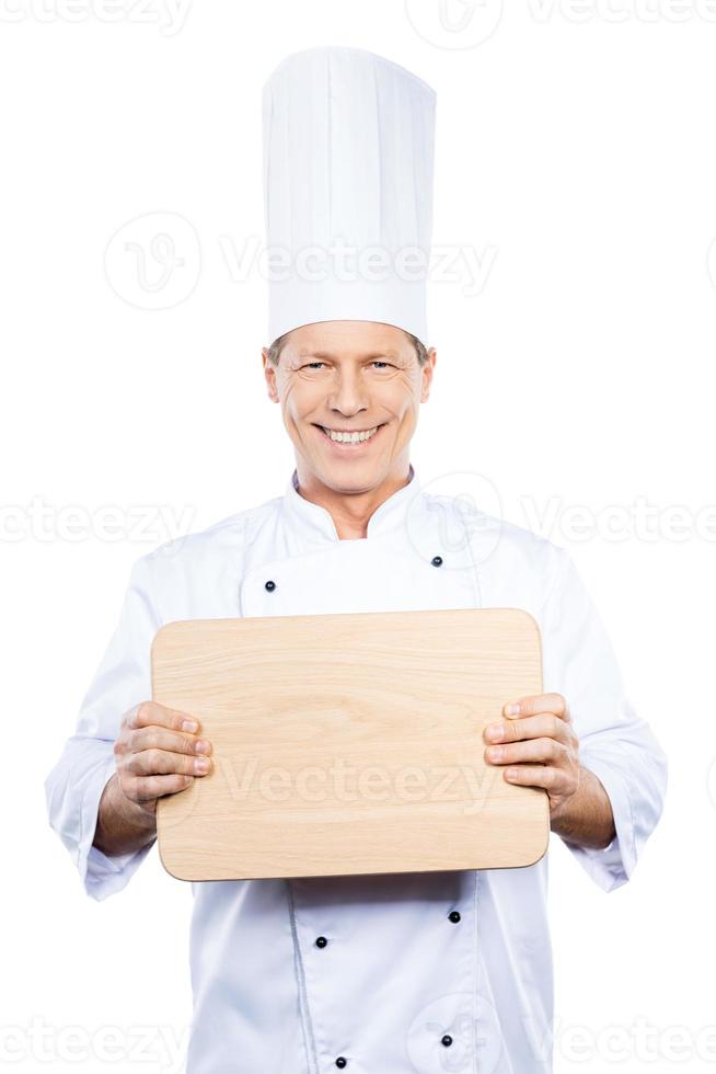 Copy space on his cutting board. Confident mature chef in white uniform holding wooden cutting board and smiling while standing against white background photo