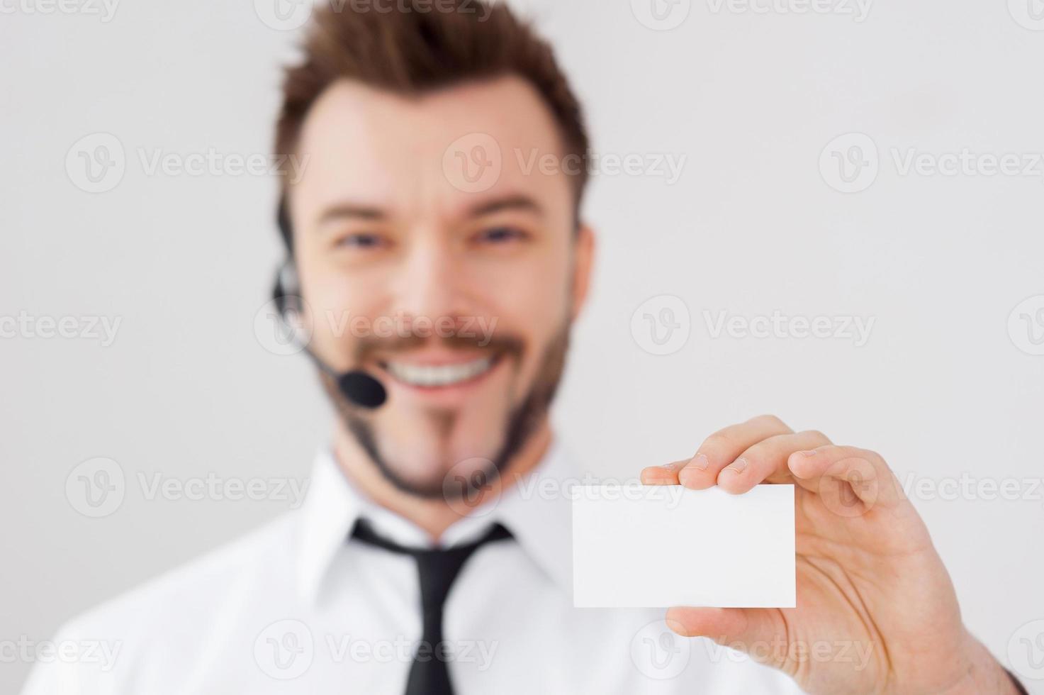 simplemente llame a este número apuesto joven en ropa formal y auriculares mostrando su tarjeta de visita y sonriendo mientras está de pie contra un fondo gris foto