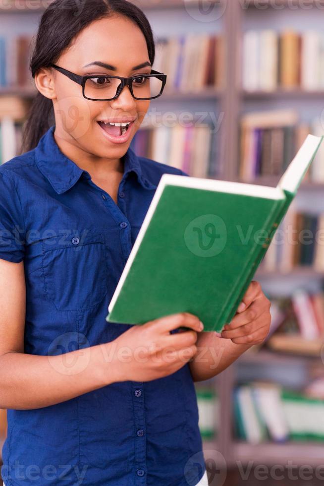 This book is so exciting Surprised African female student reading a book and keeping mouth open while standing in library photo