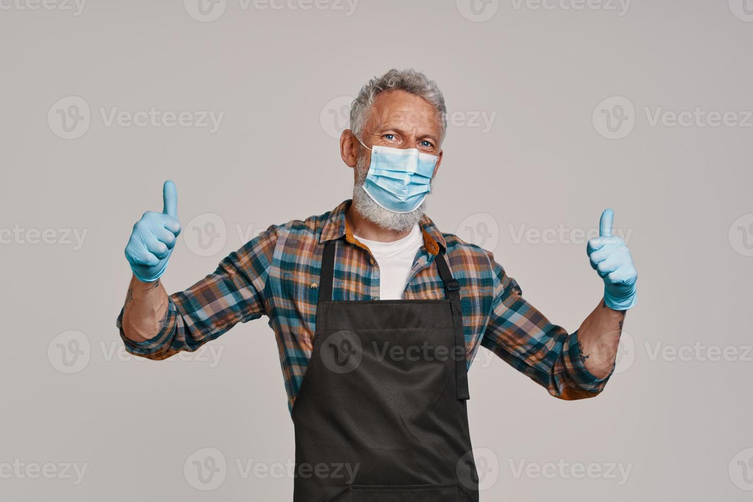 Senior man in apron and protective face mask looking at camera and keeping thumbs up photo