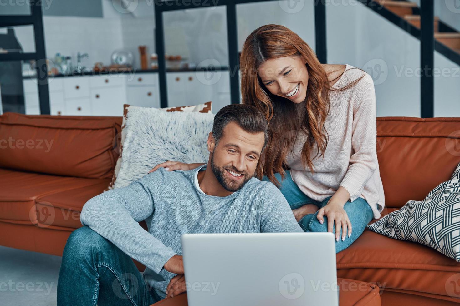 Beautiful young couple using laptop and smiling while spending time at home photo