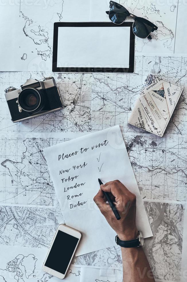 Traveling around the world. Close up top view of man writing down checklist with sunglasses, photo camera, smart phone, passport lying on map around