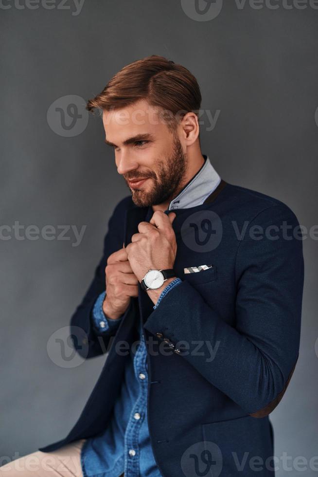 confianza y carisma. un joven apuesto mirando hacia otro lado con una sonrisa y ajustándose la chaqueta mientras se sienta en un fondo gris foto