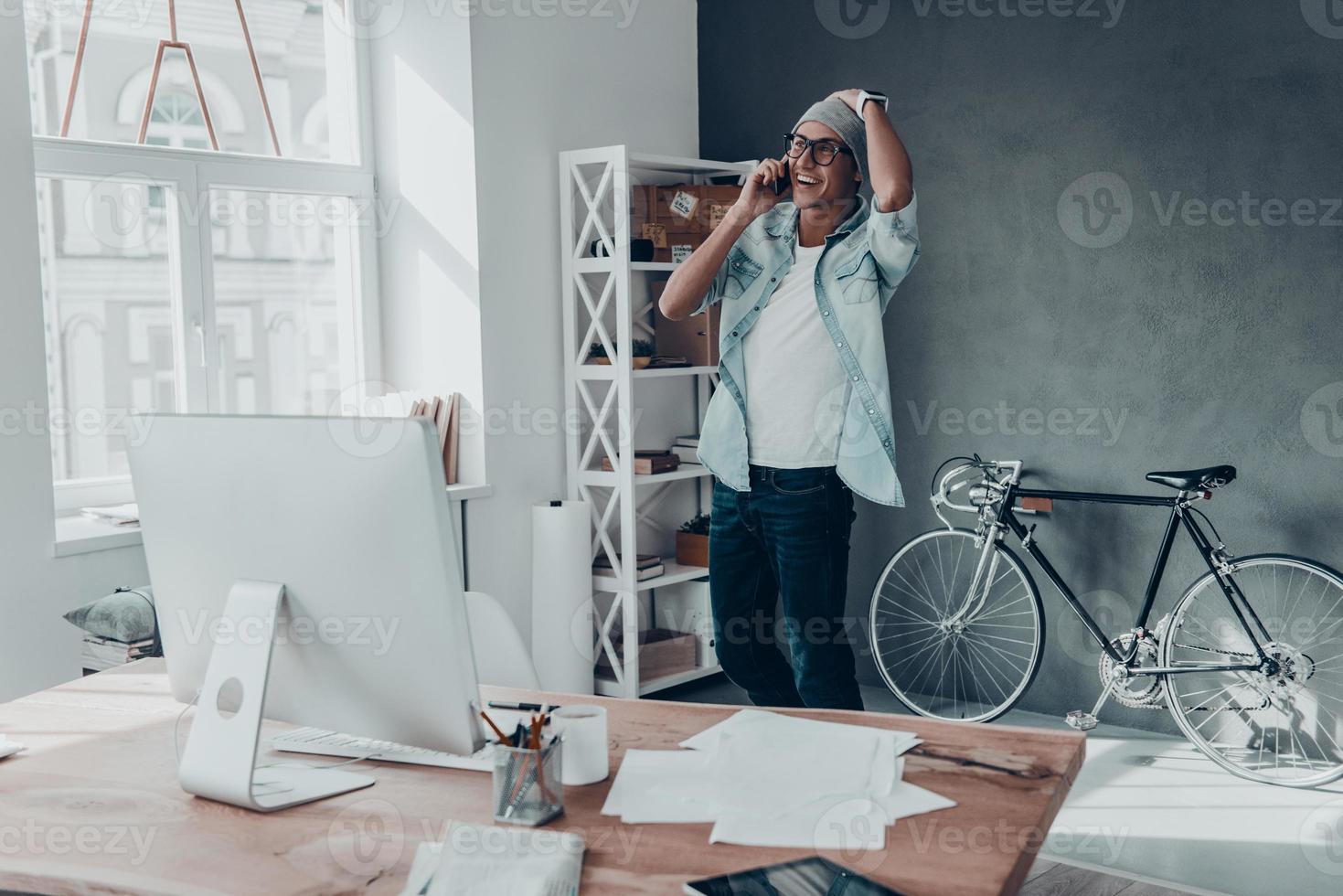 Handsome young man in eyewear talking on smart phone and smiling while standing in creative office photo