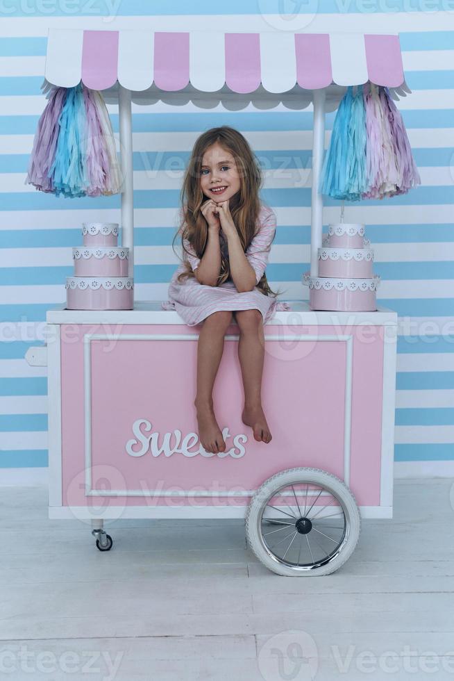 Happy little girl.  Cute little girl keeping hands clasped and smiling while sitting on the candy cart decoration photo