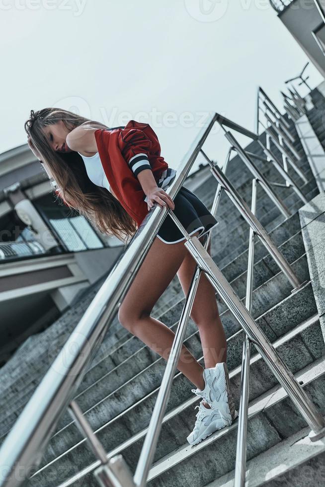 In sport style. Full length of beautiful young woman in sports uniform leaning on the railing while posing on the stairs outdoors photo