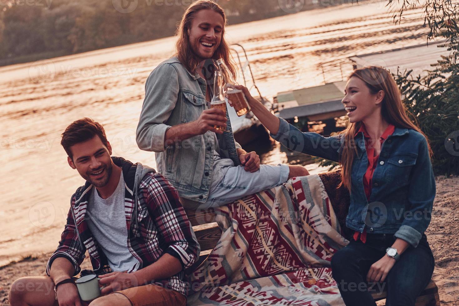 Group of young people in casual wear smiling and toasting each other while enjoying beach party near the lake photo