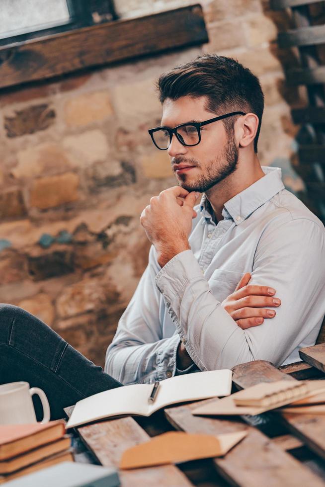 Just a moment to think.Side view of handsome thoughtful young man holding hand on chin and looking away while sitting at his working place photo