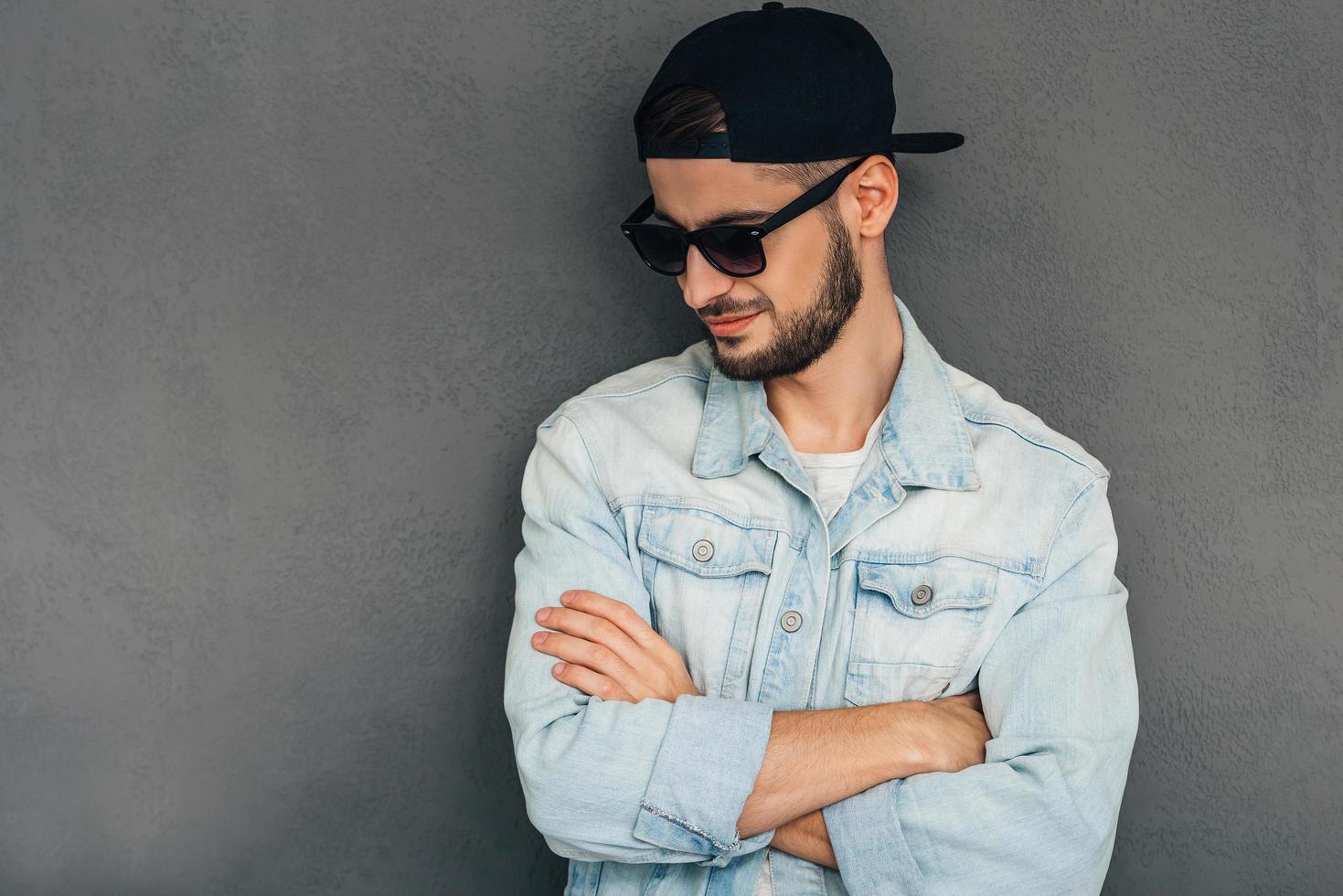 Confident in his style. Confident young man keeping arms crossed and looking down while standing against grey background photo