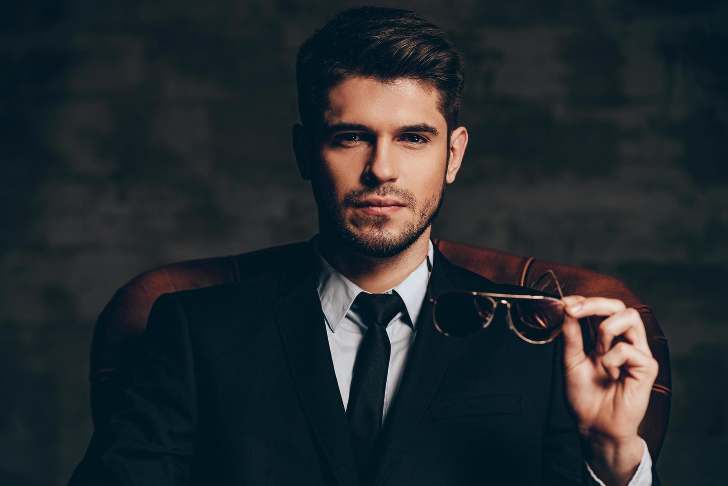 mirada impresionante.retrato de un joven apuesto con traje sosteniendo sus gafas de sol y mirando a la cámara mientras se sienta en una silla de cuero contra un fondo gris oscuro foto