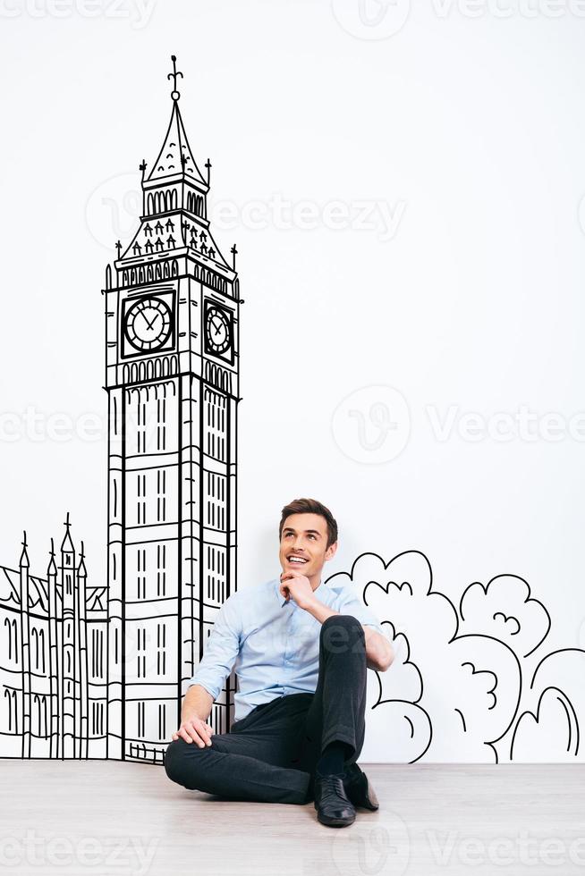 Dreaming about London. Young handsome man keeping hand on chin and looking away with smile while sitting on the floor with illustration of Big Ben at the background photo