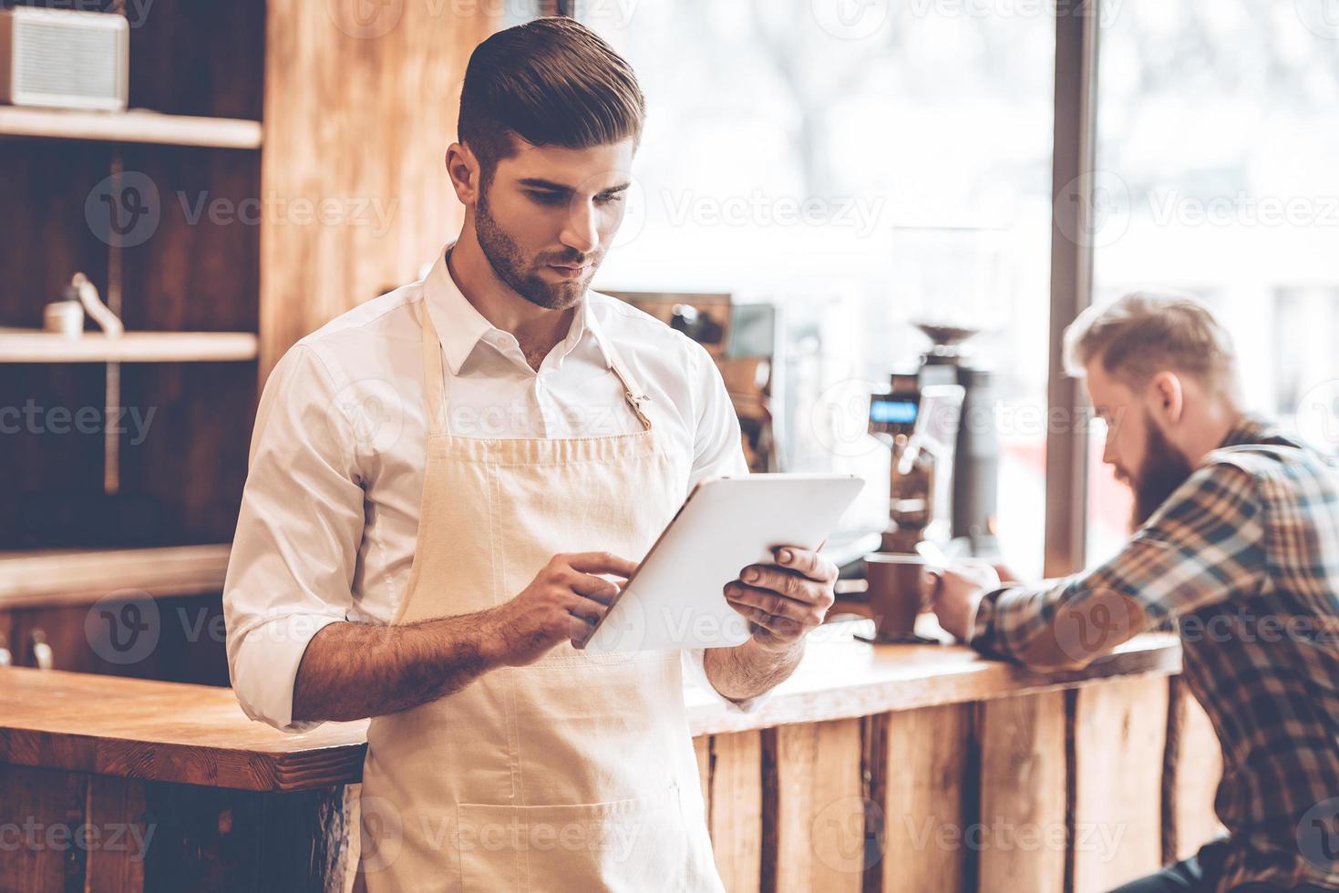 New solutions for his business. Young handsome man in apron using his digital tablet while standing at cafe with customer at background photo