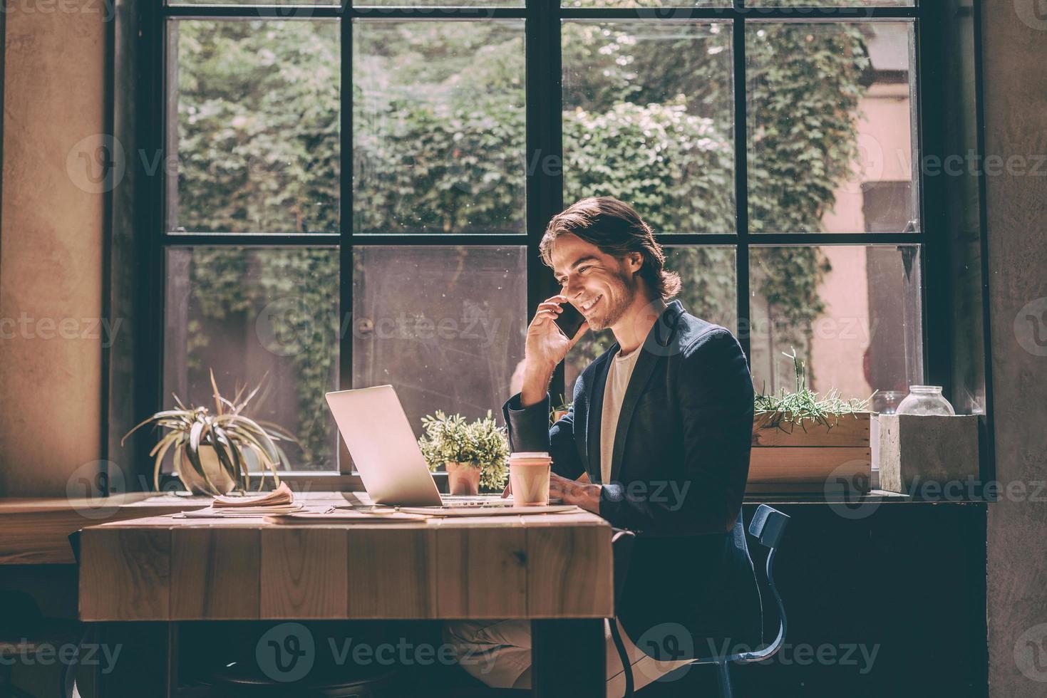 Working from cafe. Cheerful young man in smart casual wear looking at laptop and talking on the mobile phone while sitting near window in creative office or cafe photo