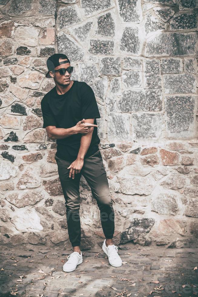 Young and cool. Full length of handsome young African man in casual clothes moving and gesturing while standing against the stoned wall outdoors photo