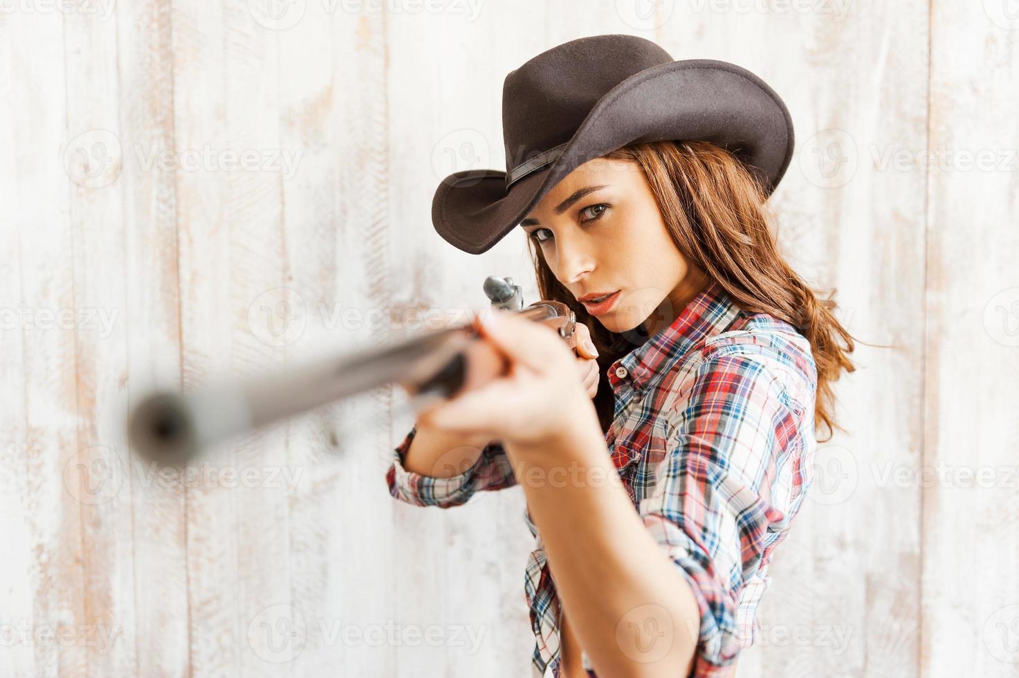 Shoot straight and look great. Beautiful young cowgirl aiming her gun at camera while standing against the wooden background photo