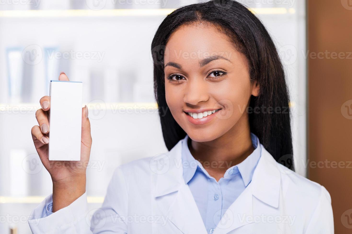 Consejo profesional. bella joven africana con bata de laboratorio sosteniendo un recipiente con un poco de medicina y sonriendo mientras está de pie en la farmacia foto